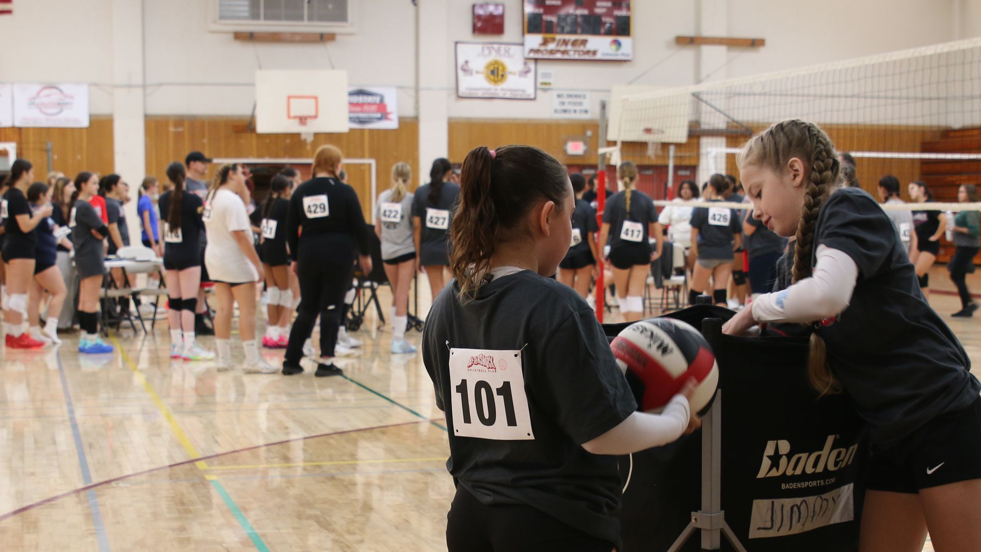 Two players in numbered tryout shirts pull volleyballs from a ball cart at the Piner High School gym, with the rest of the tryout lined up on the court behind them